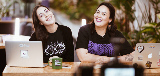 Duas mulheres sorridentes estão sentadas atrás de uma mesa. Sobre a mesa, existem dois notebooks, uma caneca e caderninhos.