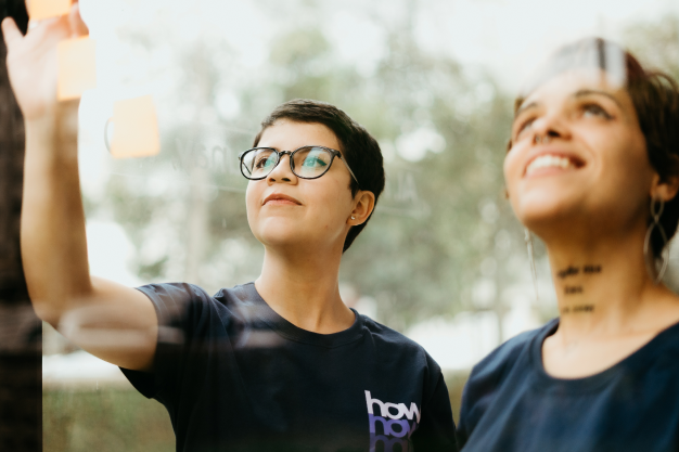 Duas mulheres estão atrás de um vidro. Elas olham para post-its de cor laranja fixados no vidro.