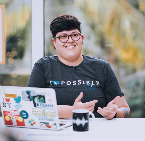 Uma mulher branca, com cabelos curtos de cor preta está sorrindo. Ela usa óculos de aro preto e está sentada atrás de uma mesa. Sobre a mesa há um notebook e uma xícara.