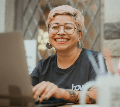 Uma mulher está sorrindo. Ela está olhando para a câmera e suas mãos estão apoiadas em um notebook.