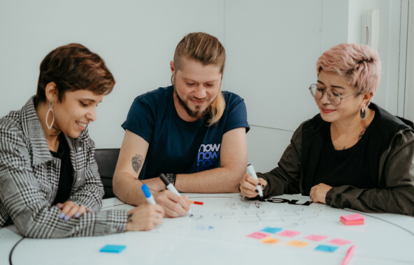 Duas mulheres e um homem sentados enquanto fazem anotações sob uma mesa com canetinhas e post-its.