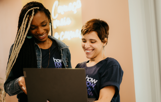 Duas mulheres estão em pé e olham para a tela de um notebook. Uma delas segura o computador, enquanto a outra aponta para no dispositivo.