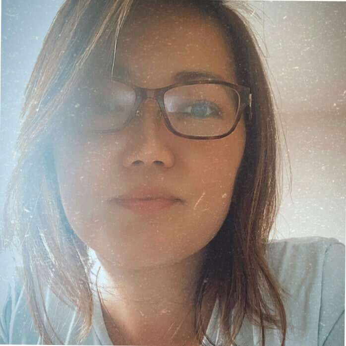 Denise é uma mulher de etnia amarela, usa óculos, tem o cabelo castanho de tamanho curto e usa uma camiseta branca.