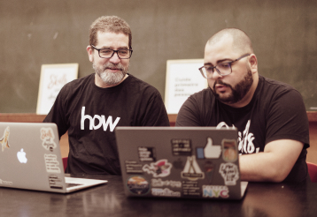 Dois homens em frente a seus notebooks. Eles olham para a tela de um dos computadores enquanto conversam.