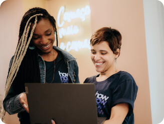 Duas mulheres estão em pé e olham para a tela de um notebook. Uma delas segura o computador, enquanto a outra aponta para no dispositivo.