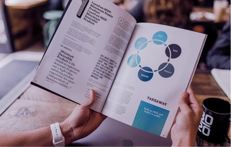 Uma mão segura uma revista, onde é possível visualizar uma ilustração de um ciclo de feedback construir, medir e aprender.