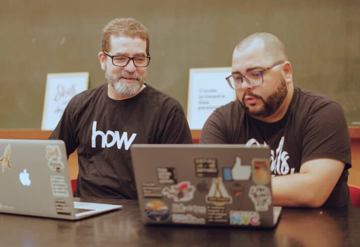 Dois homens em frente a seus notebooks. Eles olham para a tela de um dos computadores enquanto conversam.