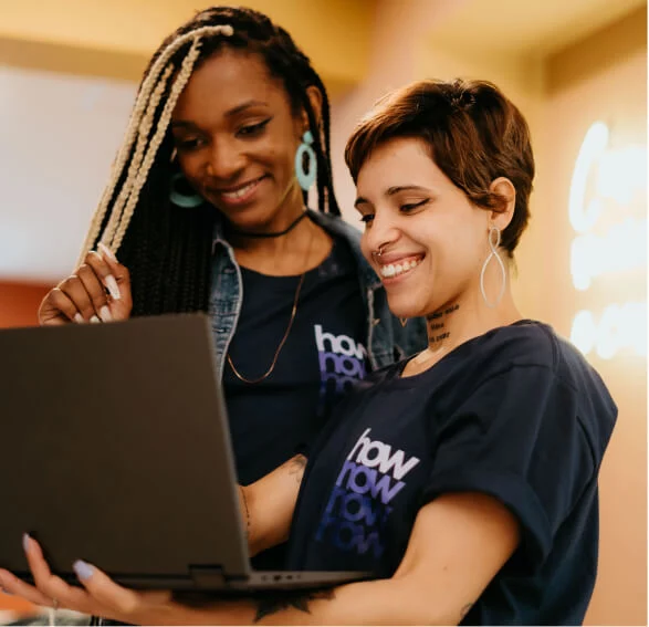 Duas mulheres estão em pé e olham para a tela de um notebook. Uma delas segura o computador. Elas sorriem.