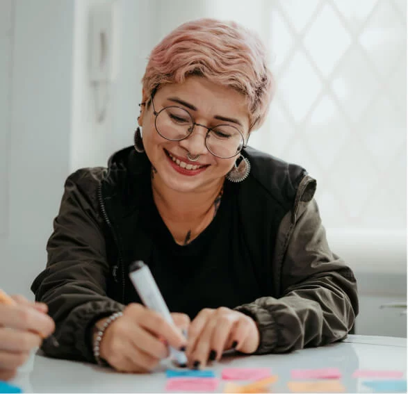Uma mulher de cabelo curto e cor rosa, está sentada e escreve com um canetão em uma nota autoadesiva. Ela está sorrindo.