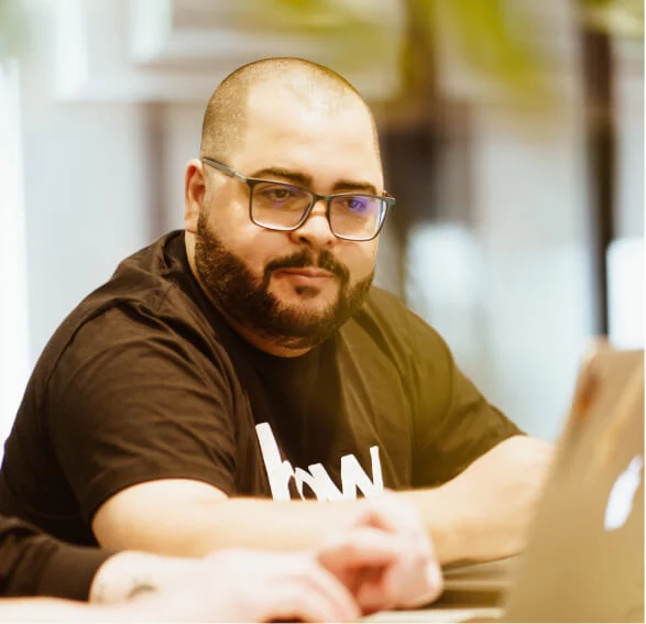 Um homem com óculos quadrado está sentando e olha para a tela de um computador. Ele possui o cabelo raspado e usa barba curta.
